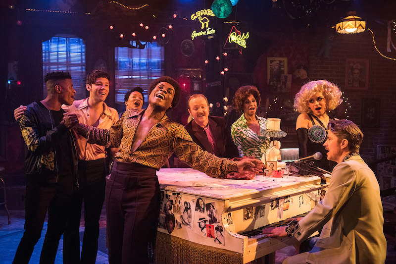 The cast sings around a white piano. They have looks of hopefulness.